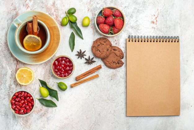 una taza de té cuencos de bayas cítricos anís estrellado y palitos de canela galletas de chocolate junto al cuaderno de crema la taza de té con canela en la mesa