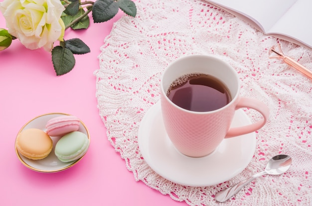 Taza de té de hierbas con macarrones; cuchara; Rosa; pluma y libro sobre fondo rosa