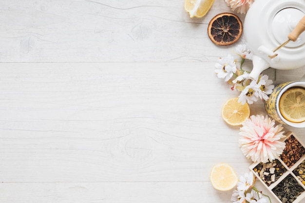 Foto gratuita una taza de té de limón; flores frescas; hierbas; hojas secas de te; tetera y rodaja de limón en mesa de madera blanca