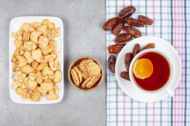 Una taza de té con limón y un montón de dátiles sobre una toalla junto a chips de galleta en un cuenco y un plato sobre fondo de mármol. Foto de alta calidad