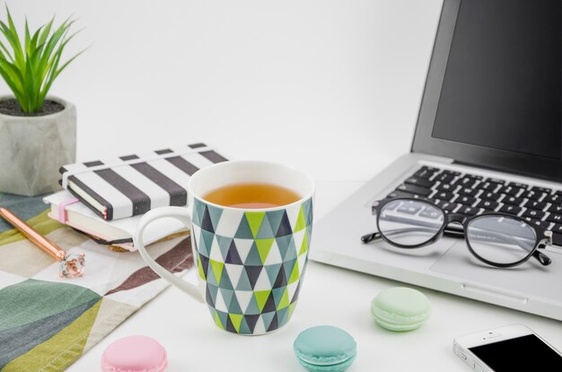 Taza de té con macarrones en la mesa de trabajo blanca con computadora portátil y teléfono móvil