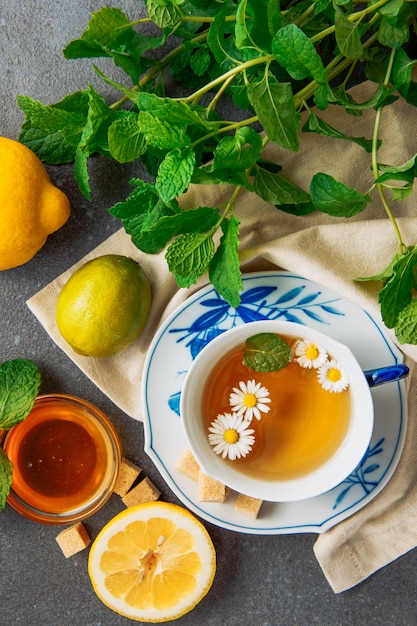 Taza de té de manzanilla en un plato con limones, terrones de azúcar morena, miel en un tazón de vidrio y hojas verdes planas sobre un fondo gris y un trozo de tela