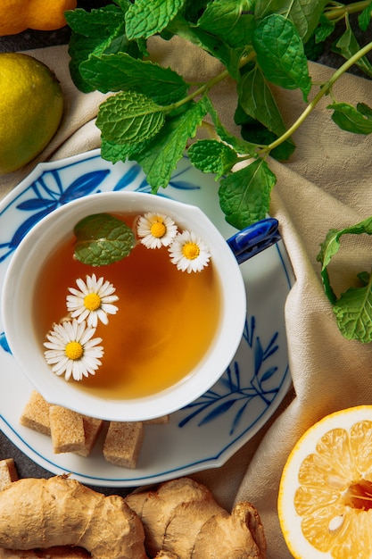 Taza de té de manzanilla con rodajas de limón, jengibre, terrones de azúcar morena y hojas verdes en un platillo sobre fondo de tela, vista de ángulo alto.