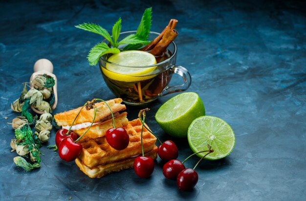 Taza de té con menta, canela, hierbas secas, cereza, lima sobre superficie azul sucio, vista de ángulo alto.