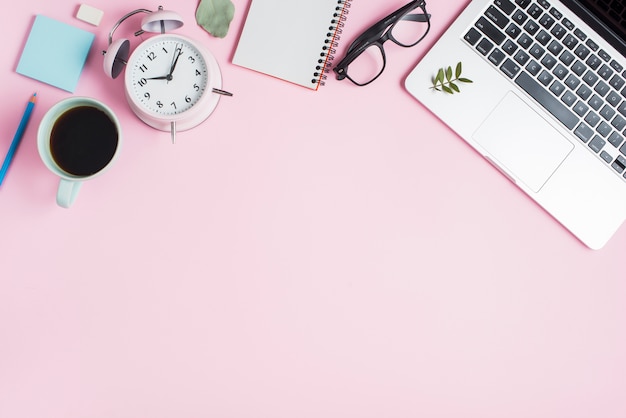 Taza de té negro; despertador; bloc de notas espiral anteojos y laptop sobre fondo rosa