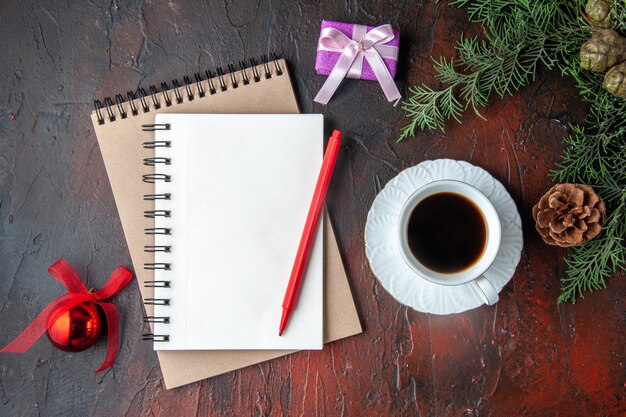 Una taza de té negro, ramas de abeto, accesorios de decoración y regalo junto al cuaderno con bolígrafo sobre fondo oscuro