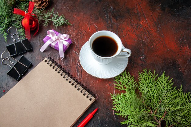 Una taza de té negro, ramas de abeto, accesorios de decoración y regalo junto al cuaderno con bolígrafo sobre fondo oscuro