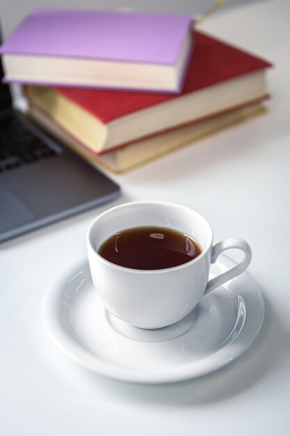 Taza de té con platillo sobre la mesa cerca de libros y portátil