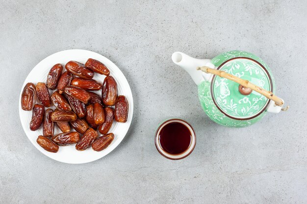 Una taza de té y una tetera adornada junto a un plato de dátiles frescos sobre fondo de mármol. Ilustración de alta calidad