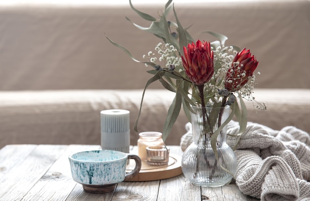 Foto gratuita taza, velas, jarrón con flores de protea y un elemento de punto en la habitación sobre un fondo difuminado.