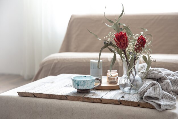 Taza, velas, jarrón con flores de protea y un elemento de punto en la habitación sobre un fondo difuminado.