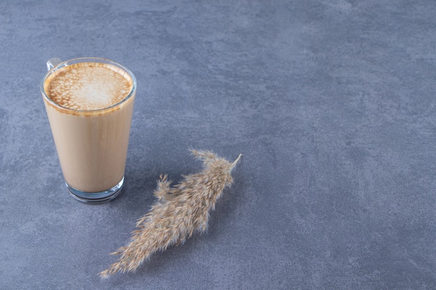 Taza de vidrio de café con leche junto a la hierba de la pampa, sobre la mesa azul.