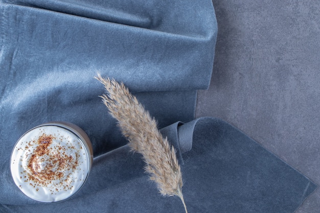 Taza de vidrio de café con leche en un trozo de tela junto a la hierba de la pampa, sobre la mesa azul.