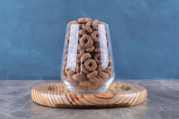 Una taza de vidrio llena de anillos de cereal de chocolate sobre una tabla de madera.