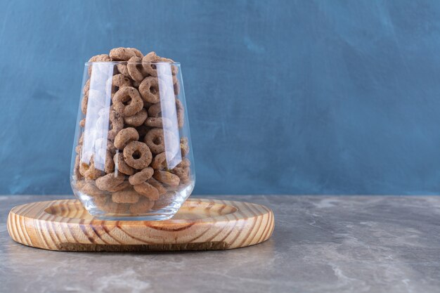 Una taza de vidrio llena de anillos de cereal de chocolate sobre una tabla de madera.