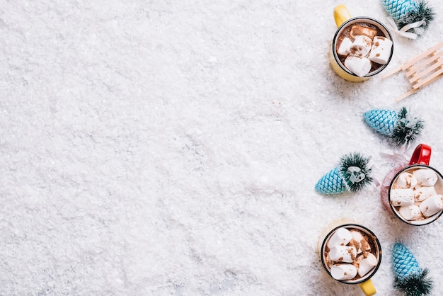 Tazas con malvaviscos y bebidas cerca de juguetes navideños entre nieve