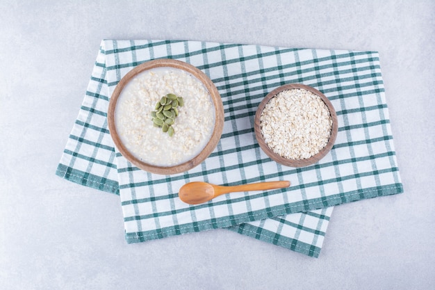 Un tazón de avena y un tazón de avena cubierto con pepitas, junto a una cuchara sobre una toalla sobre la superficie de mármol