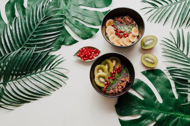 Foto gratuita tazón de batido de aguacate cubierto con chía, granola, kiwi y espinacas. vista aérea, superior, endecha plana. desayuno saludable. hojas tropicales.