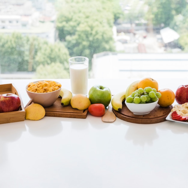 Tazón de copo de maíz y frutas coloridas con vaso de leche en mesa blanca cerca de la ventana