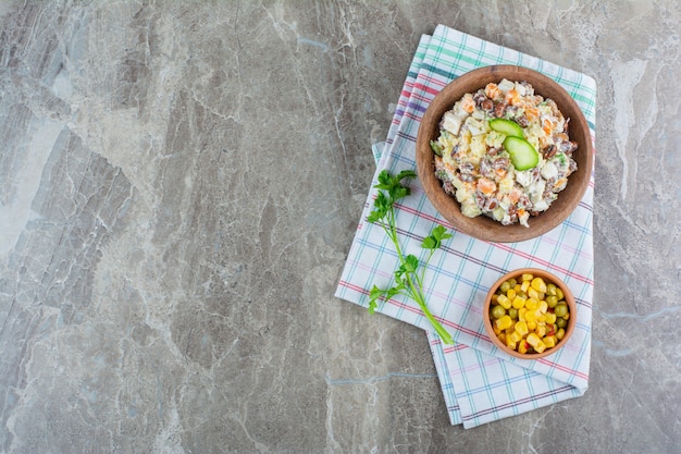 Un tazón de ensalada de verduras junto a una ensalada de maíz en un tazón sobre un paño de cocina sobre mármol.