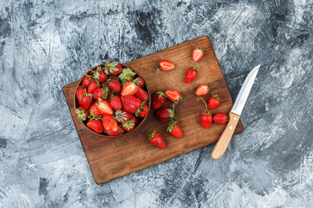 Un tazón de fresas y un cuchillo sobre una tabla de cortar de madera sobre un fondo de mármol azul oscuro. endecha plana.