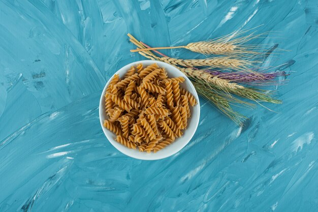 Tazón de fuente blanco de pasta fusilli seca con espigas de trigo sobre superficie azul