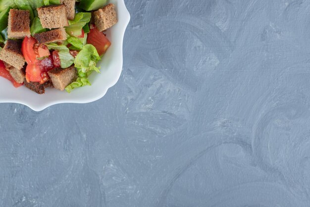 Tazón de fuente de ensalada de verduras con tomate, pepino, lechuga y corteza seca sobre mesa de mármol.