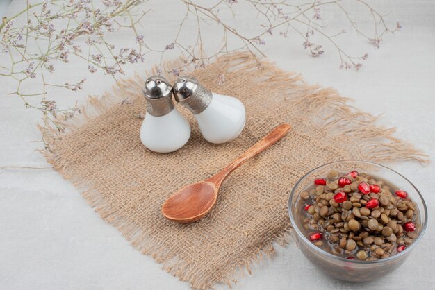 Tazón de fuente de frijoles hervidos con semillas de granada sobre superficie blanca con sal.