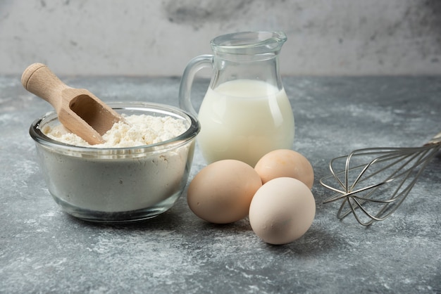 Tazón de fuente de harina, huevos y utensilios de cocina en la mesa de mármol.