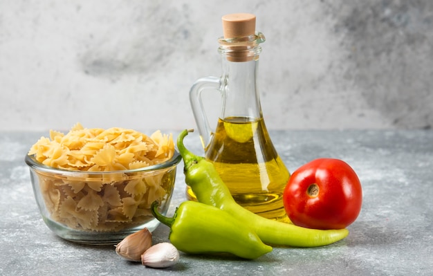 Tazón de fuente de pasta farfalle cruda con botella de aceite de oliva y verduras en la mesa de mármol.