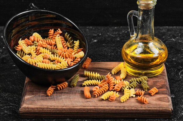 Tazón de fuente de pasta fusilli colorida y botella de aceite en la mesa oscura.