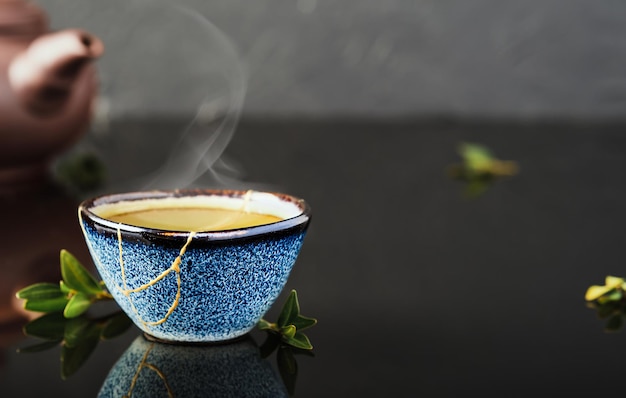 Tazón de fuente de té verde japonés, las hojas de té se encuentran al lado de la taza. Enfoque selectivo en la taza. El vapor sube por encima del recipiente. Taza azul de cerámica recuperada, segunda vida de las cosas, reciclaje o kintsugi