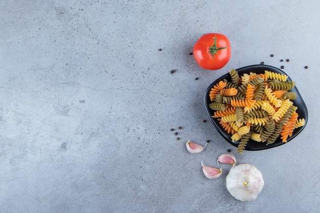 Un tazón negro lleno de macarrones multicolores con tomate rojo fresco y ajo sobre un fondo de piedra.