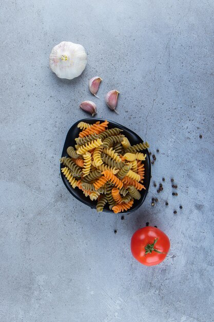 Un tazón negro lleno de macarrones multicolores con tomate rojo fresco y ajo sobre una superficie de piedra.