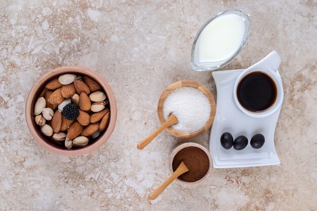 Un tazón de nueces variadas, tazones pequeños de leche, café molido, azúcar y una taza de café.