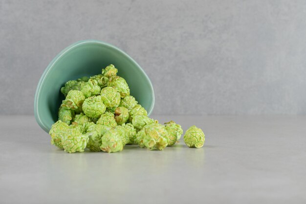 Tazón pequeño caído, derramando palomitas de maíz confitadas verdes sobre fondo de mármol.