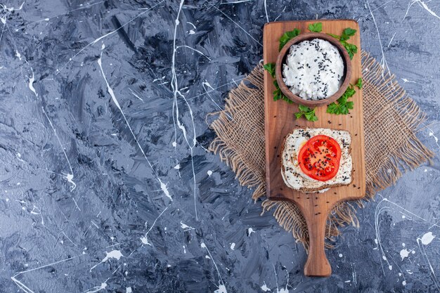Un tazón de queso junto a los tomates en rodajas en un pan de queso sobre una tabla de cortar sobre una servilleta de arpillera, en el azul.