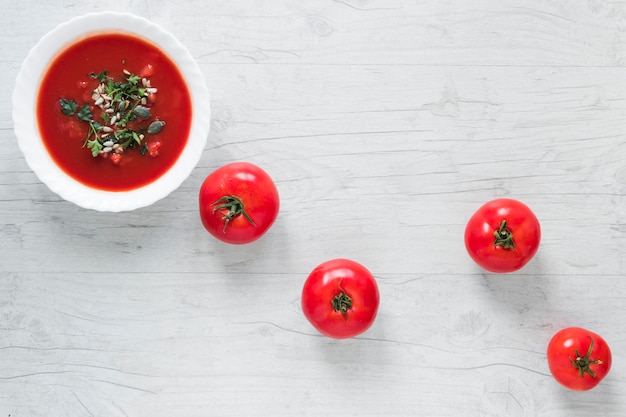 Un tazón de sopa de tomate fresco en un tazón de cerámica blanco adornado con hierbas y tomates maduros en mesa de madera