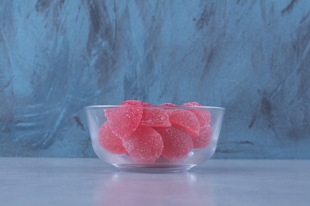 Foto gratuita un tazón de vidrio lleno de caramelos de gelatina azucarados rojos sobre la mesa gris.