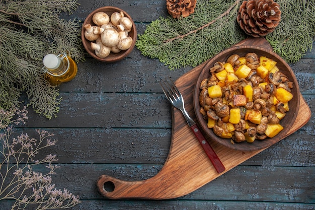 Tazón de vista superior del tazón de comida de champiñones y patatas en la tabla de cortar junto al tenedor debajo de la botella de aceite tazón de setas blancas y ramas de abeto