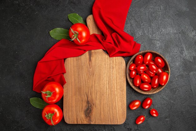 Tazón de vista superior con tomates cherry, toalla roja, una tabla de cortar y tomates rojos sobre fondo oscuro