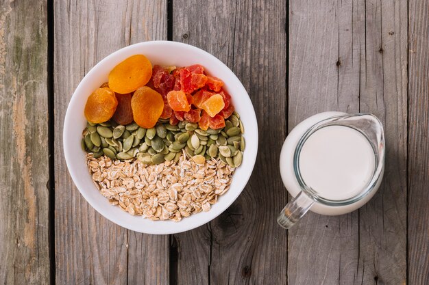 Tazones de fuente de muesli, semillas de calabaza y frutas secas en el tazón con jarra de leche en la mesa de madera rústica