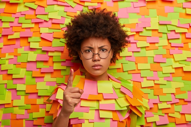 Foto gratuita ¡te dije! mujer de piel oscura enojada y disgustada advierte a alguien, levanta el dedo índice con expresión estricta, muestra un gesto de amonestación, saca la cabeza, rodeada de notas adhesivas de papel de colores