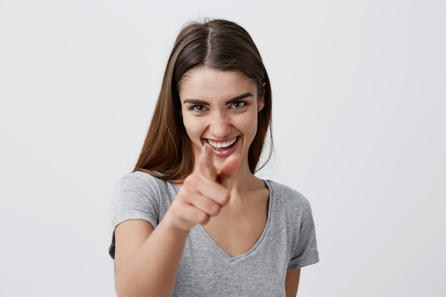 Te encuentro Primer plano de una joven y bella muchacha caucásica de cabello oscuro y graciosa con cabello de registro en camisa gris sonriendo con dientes, señalando con las manos, divirtiéndose durante la sesión fotográfica familiar.