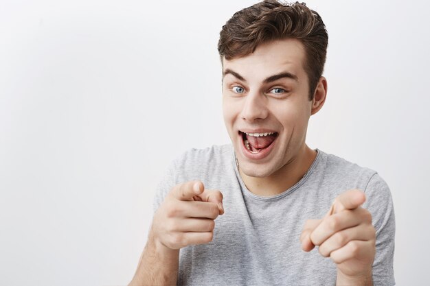 ¡Te escojo a ti! El hombre emocional positivo con cabello oscuro te mira con alegría y te señala con los dedos índices. Guapo joven apuesto divirtiéndose ondoors, gestos y expresiones faciales.