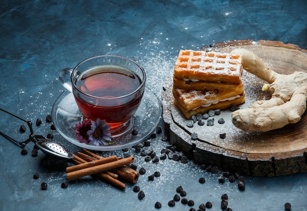 Té con gofres, choco chips, especias, colador en una taza en azul grunge y superficie de tablero de madera, vista de ángulo alto.