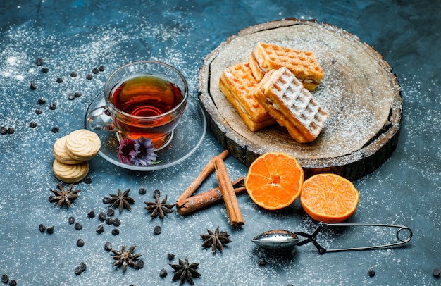 Té con gofres, galletas, especias, choco chips, colador, naranja en una taza en la superficie de tablero azul y madera, vista de ángulo alto.