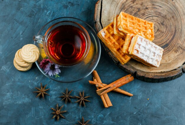 Té con gofres, galletas, palitos de canela, especias en una taza sobre una superficie de tablero de madera y azul sucio, plano.