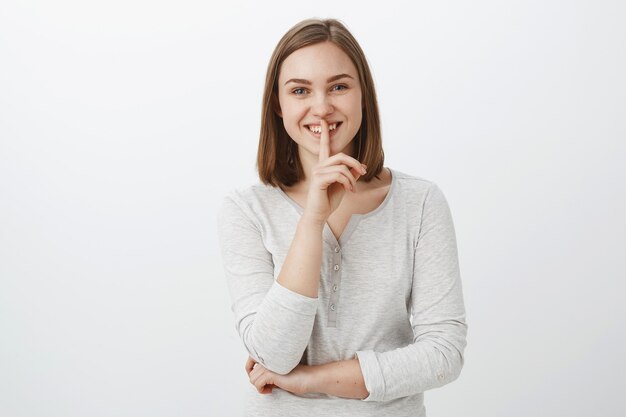 Te he dicho algo pero shh. Retrato de mujer joven encantadora entusiasta de aspecto amistoso con corte de pelo marrón corto mostrando gesto de silencio con el dedo índice sobre la boca sonriente escondiendo secreto