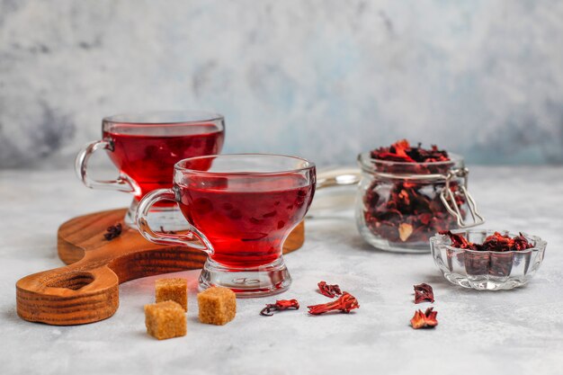 Té de hibisco candente en una taza de vidrio sobre concreto con pétalos de hibisco secos